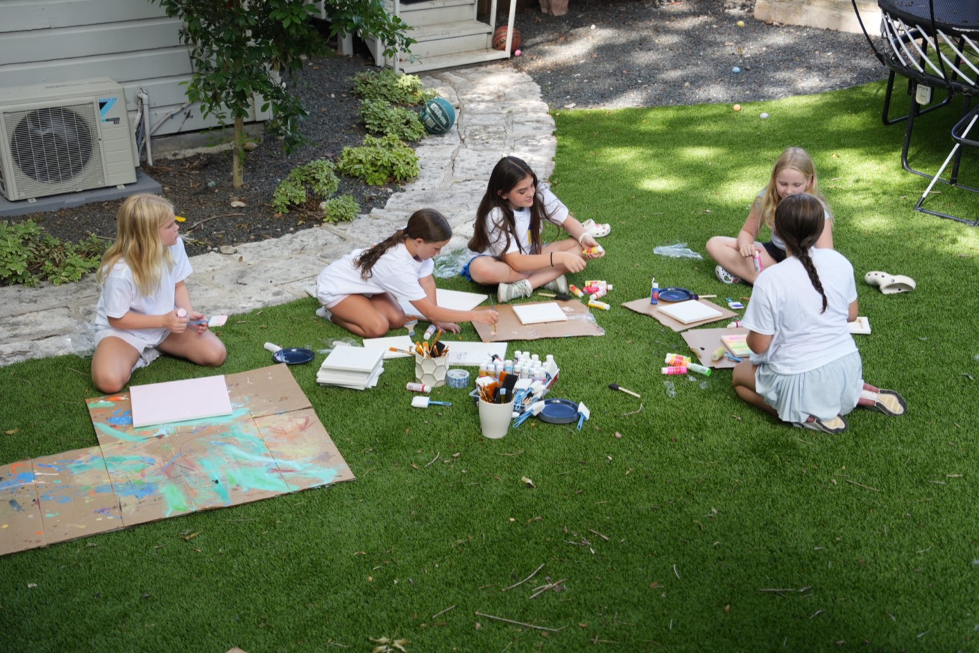 Five campers sitting on the lawn painting canvases, surrounded by brushes and paint.